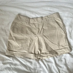 Beige bomullsshorts med fickor - Snygga beige shorts i bomull med klassisk design. De har två sidofickor fram, två bakfickor med knapp och hällor för bälte. Stängs med dragkedja och knapp framtill. Perfekta för varma dagar och enkel att matcha med allt