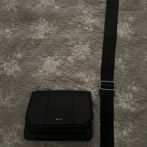 Svart axelväska från NA-KD - Snygg svart axelväska från NA-KD med stilren design och justerbart brett axelband. Väskan är tillverkad i skinnimitation och har en minimalistisk look med diskret logga framtill. Perfekt för att bära dina viktigaste grejer. Pris kan diskuteras !!