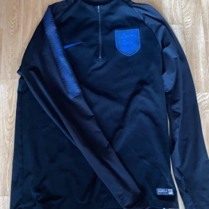 England Nike Dri-Fit långärmad tröja S - Säljer en svart långärmad England fotbollströja från Nike med blått landslagsmärke och Nike-logga. Tröjan har halv dragkedja och är tillverkad i Dri-Fit-material som andas. Perfekt för träning eller match.
