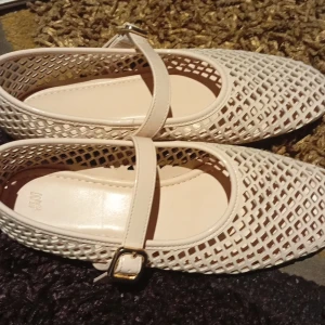 Beige ballerinaskor med spänne från H&M - Snygga beige ballerinaskor från H&M med coolt rutmönster i syntetmaterial. Skorna har en klassisk Mary Jane-rem med guldfärgat spänne och platt sula. Perfekta för dig som gillar en luftig och trendig stil med en twist.