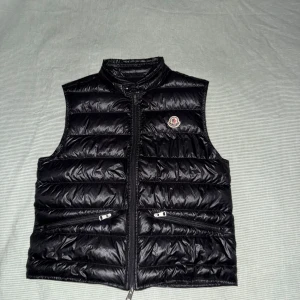 Svart dunväst från Moncler - Svart dunväst från Moncler med quiltad design och hög krage. Västen har två dragkedjefickor framtill och stängs med dragkedja. Klassisk Moncler-logga på bröstet. Perfekt för lager-på-lager och snygg till streetwear. Size 2
