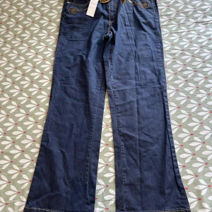 Blå wide jeans med guldbroderi DNY - Snygga blå jeans från DNY med breda ben och coola guldbroderier samt detaljer i guld. Byxorna har en midwaist passform och är tillverkade i bomull med lite stretch för extra komfort. Perfekta för dig som vill sticka ut med unika detaljer.