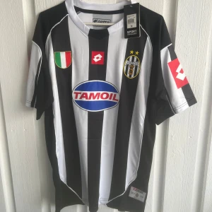 Juventus Del Piero #10 matchtröja Lotto - Juventus fotbollströja med Del Piero #10 på ryggen. Klassisk svartvit randig design, officiella klubbmärket och Tamoil-sponsor på bröstet. Korta ärmar, broderade detaljer och tillverkad i lätt polyester. Perfekt för fans av italiensk fotboll och Del Piero.