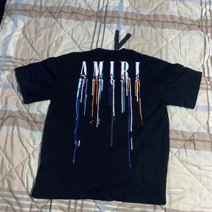 Svart AMIRI t-shirt med färgstänk - Svart t-shirt från AMIRI med coolt tryck av droppande färg och stor logga både fram och bak. T-shirten har rund hals, korta ärmar och är gjord i mjuk bomull. Perfekt för dig som vill sticka ut med streetstyle-vibe.