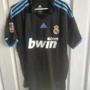 Real Madrid Ronaldo 9 svart Adidas tröja - Snygg svart Real Madrid fotbollströja från Adidas med ljusblå detaljer och klassiska tre ränder på axlarna. Tryck med Ronaldo 9 på ryggen och Bwin-logga på bröstet. Officiellt klubbmärke och LFP-patch på ärmen. Tillverkad i lätt och ventilerande material.