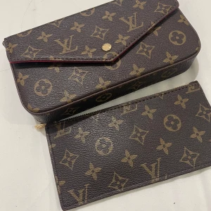  axelväska - Säljer en klassisk axelväska i brunt skinn med ikoniskt monogrammönster och guldfärgade detaljer. Väskan har röd insida, avtagbar innerficka med dragkedja och medföljande guldfärgad kedja. Perfekt för dig som vill ha en lyxig och tidlös accessoar.