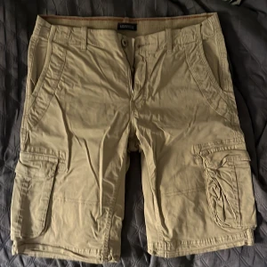 Lindbergh Cargo Shorts - Beige - Storlek S - Snygga beige cargoshorts från Lindbergh med flera praktiska fickor på sidorna och bak. Klassisk passform med bälteshällor och knappstängning. Perfekta för varma dagar och chill stil. Materialet känns lätt och skönt mot huden. Äkta vara köpt från Lindbergh.