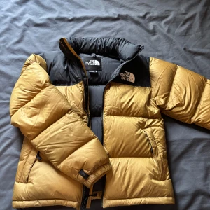 The North Face gul och svart dunjacka XS - Säljer en ikonisk pufferjacka från The North Face i storlek XS. Jackan är gul med svarta detaljer, har hög krage, dragkedja och två sidofickor. Perfekt för kalla dagar med sin dunfyllda insida och klassiska logga på bröstet och ryggen.