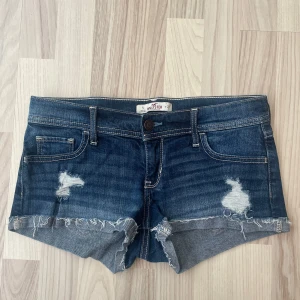 Mörkblå jeansshorts med slitningar från Hollister - Snygga mörkblå jeansshorts från Hollister med slitna detaljer och fransig kant. Klassisk femficksmodell med låg midja och coola slitningar framtill och på bakfickorna. Perfekta för sommaren och ger en avslappnad look💞💞