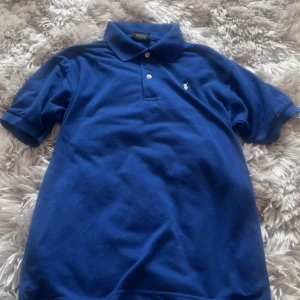 Blå pikétröja från Polo  - Klassisk blå pikétröja från Polo Sport med vit broderad logga på bröstet. Tröjan har krage, två knappar och korta ärmar. Tillverkad i 100% bomull för en skön och luftig känsla. Perfekt för en sportig och stilren look.