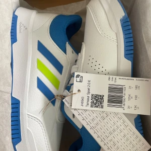 Adidas Tensaur Sport 2.0 sneakers - Helt nya Fräscha Adidas Tensaur Sport 2.0 sneakers i vitt med blå och limegröna detaljer. Skorna har klassiska tre ränder på sidan, vit ovandel i syntet och blå meshfoder. Snygg design med snörning och slitstark sula som inte lämnar märken.