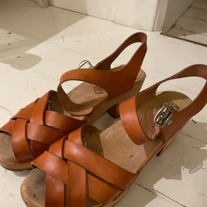 Bruna träskosandaler från Complement - Snygga bruna sandaler i klassisk träskostil från Complement. Skorna har flätade remmar i skinn, justerbart spänne i silver och en stadig träsula med låg klack. Perfekta för dig som gillar retrovibbar och naturliga material.