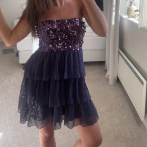 Mörklila paljettklänning med volanger - Säljer en mörkblå axelbandslös kortklänning med glittriga paljetter upptill och flera lager volanger i kjolen. köpt secondhand 