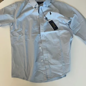 Ljusblå skjorta från Ralph Lauren - Klassisk ljusblå skjorta från Ralph Lauren med button-down krage och broderad logga på bröstet. Skjortan har lång ärm, knappar framtill och är tillverkad i bomull. Perfekt för en clean och stilren look.