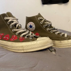 Converse x CDG Play Chuck Taylor 42,5 - Gröna Converse Chuck Taylor All Star high-tops i samarbete med Comme des Garçons Play. Skorna har vita snören, vit sula och röda hjärtan med ögon längs sidan. Klassisk rund tå och canvasmaterial. Perfekt för dig som vill sticka ut med en ikonisk sneaker.