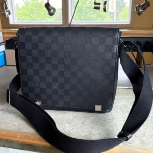 Louis Vuitton Damier Graphite axelväska - Snygg axelväska från Louis Vuitton i klassiska Damier Graphite-mönstret. Väskan är svart med rutigt mönster och har bred, justerbar axelrem. Tillverkad i slitstarkt skinn och textil, perfekt för dig som vill ha en stilren och praktisk väska med lyxig känsla.