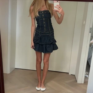 Mörkblå jeansklänning - Supersöt mörkblå jeansklänning med volangkjol och knappar framtill. Axelbandslös modell med markerad midja och coola sömdetaljer.