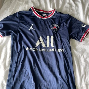 PSG matchtröja blå Jordan x Nike - Snygg PSG fotbollströja i mörkblått med vita och röda detaljer på krage och ärmslut. Tröjan har klubbmärke, Jordan-logga och sponsortryck på bröstet. Tillverkad i lätt och ventilerande polyester, perfekt för fotboll eller streetwear.