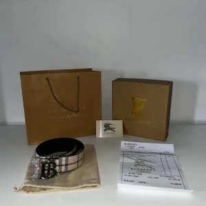 Burberry rutigt skärp med B-spänne - Snyggt Burberry-bälte i klassiskt beige och brunt rutig design med stort B-spänne i metall. Bältet är gjort i skinn och har den ikoniska Burberry-looken. Perfekt accessoar för att lyfta din outfit med en lyxig touch.