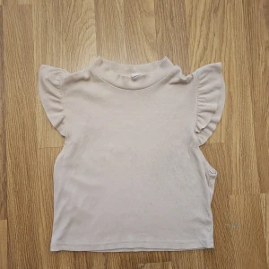 Beige ribbad ärmlös topp med volang - Supersöt beige topp med ribbad struktur och volangdetaljer på axlarna. Hög hals och ärmlös design ger en clean och trendig look. Perfekt att matcha med jeans eller kjol för en chill stil.