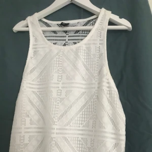 Vit långklänning med spetsdetaljer - Vit långklänning med ärmlös design och snygga spetsdetaljer upptill. Klänningen har ett geometriskt mönster i spetsen och ett skirt, flowy tyg nedtill som ger en luftig känsla. Perfekt för sommarens alla tillfällen när du vill känna dig extra fin.