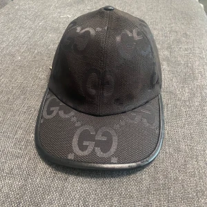 Svart keps från Gucci med GG-mönster - Snygg svart keps från Gucci med ikoniskt GG-mönster ton-i-ton över hela kepsen. Bågen är böjd och kepsen har justerbart spänne bak. Materialet är slitstarkt och har en lyxig känsla, perfekt för dig som gillar streetstyle och designeraccessoarer. Kan gå ner vid pris vid snabbäffär och därmed att jag inte säljer boxen. 