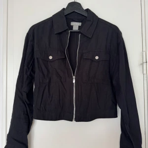 Svart croppad linnejacka från Lindex - Svart croppad linnejacka från Lindex med klassisk krage, silverfärgad dragkedja och två bröstfickor med knappar. Jackan har lång ärm och en clean, stilren look som passar perfekt till dina favoritjeans eller kjol.