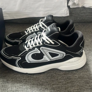 Svarta Dior B30 sneakers strl 41 - Säljer ett par svarta Dior B30 sneakers med vita detaljer och snörning. Skorna har en chunky vit sula, mesh och skinnpaneler samt den ikoniska CD-loggan på sidan. Riktigt snygga och trendiga sneakers med sportig vibe. Levereras med originalkartong.