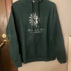 Mörkgrön hoodie från Madlady - Säljer en mörkgrön hoodie med snyggt månfas- och soltryck både fram och bak. Tröjan har huva med snörning och en stor magficka. 