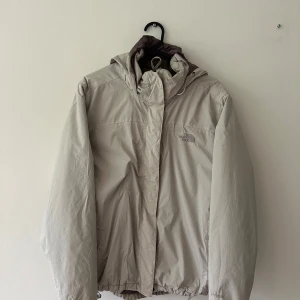 Beige vindjacka från The North Face - Snygg beige vindjacka från The North Face med huva och dragsko nertill. Jackan har diskret logga på bröstet och är tillverkad i slitstarkt syntetmaterial. Perfekt för blåsiga dagar och enkel att matcha med olika outfits.