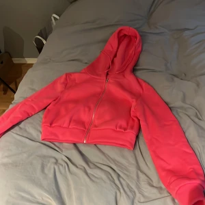 Croppad rosa hoodie med dragkedja - Snygg croppad hoodie i stark rosa färg med dragkedja framtill och huva. Tröjan har långa ärmar och bred mudd vid midjan för en modern look. Perfekt för dig som gillar streetstyle och vill ha något bekvämt men ändå trendigt.