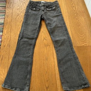 Grå bootcut jeans från Gina Tricot - Snygga grå jeans från Gina Tricot i bootcut-modell med coola fickdetaljer och knappar framtill. Jeansen har en klassisk femficksdesign, bälteshällor och är gjorda i mjukt jeanstyg. Perfekt för dig som gillar en avslappnad men trendig look. Jeansen är i gott skick och i storlek 152.