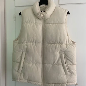 Beige dunväst från Lindex - Säljer en beige dunväst från Lindex i storlek M. Västen har hög krage, två snedställda fickor och dragkedja framtill. Den har en clean, oversized look som är perfekt för lager-på-lager. Vet ej frakt kostnaden.
