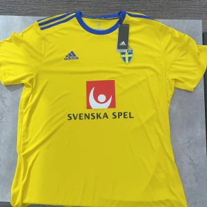 Svenska landslagströja Adidas XL - Gul fotbollströja från Adidas i storlek XL, officiell landslagströja för Sverige med blå detaljer och blått krage. Tryck med Svenska Spel på bröstet och Sveriges landslagsemblem. Tillverkad i lätt och ventilerande Climalite-material.