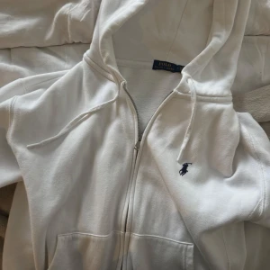 Vit hoodie från Polo Ralph Lauren - Säljer en klassisk vit hoodie från Polo Ralph Lauren i bomull. Tröjan har dragkedja framtill, huva med snörning och den ikoniska broderade loggan på bröstet. Perfekt för en chill och stilren look.