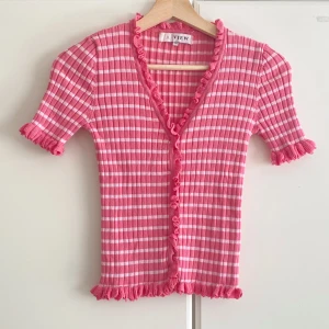 Rosa och vit randig topp från A View - Supersöt rosa och vit randig topp från A View med korta ärmar, v-ringning och volangdetaljer längs kanterna. Toppen är ribbad och har en croppad känsla med figurnära passform. Perfekt för dig som gillar färg och lekfull stil.