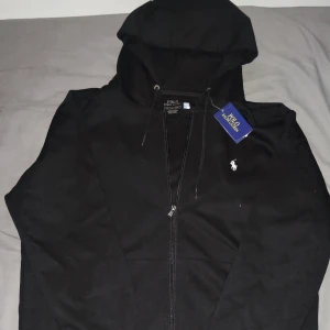 Svart hoodie från Polo Ralph Lauren - Svart hoodie från Polo Ralph Lauren med klassisk vit logga på bröstet. Tröjan har huva med snörning, dragkedja framtill och två fickor. Tillverkad i mjuk bomull, perfekt för en chill och stilren look.