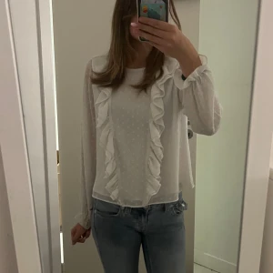 Vit blus med volangdetaljer - Supersöt vit blus med diskreta prickar och stora volanger framtill. Långärmad modell med lätt transparent tyg och rund halsringning. 