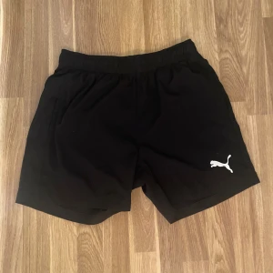 Svarta träningsshorts från Puma - Svarta shorts från Puma med elastisk midja och klassisk vit logga på ena benet. Perfekta för träning eller chill, med enkel och sportig design. Lätta och bekväma att bära, passar dig som gillar en clean look.