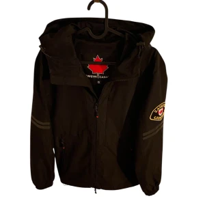 Svart vindjacka från Zavetti Canada - Svart vindjacka från Zavetti Canada med huva, dragkedja framtill och broderad logga på ärmen. Jackan har elastiska muddar, två fickor med dragkedja och reflekterande ränder på ärmen. Perfekt för dig som gillar sportig och clean stil.