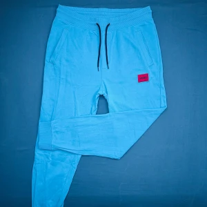 Blå mjukisbyxor från Hugo - Snygga ljusblå mjukisbyxor från Hugo med svart snörning i midjan och ribbade muddar vid benslut. Byxorna har två sidofickor, en bakficka och en röd Hugo-logga framtill. Perfekta för chill eller streetwear.