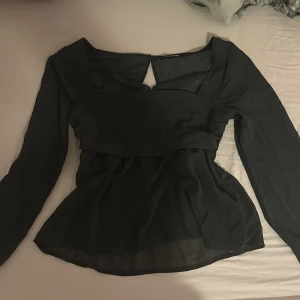 Mörk grå blus med transparenta ärmar - Säljer en svart blus i tunn polyester med långa transparenta ärmar och smockad midja. Blusen har en snygg detalj med knapp och öppning i nacken samt en lätt utsvängd passform nedtill. Perfekt för dig som gillar stilrena och trendiga plagg. Är helt oanvänd utan prislapp. 