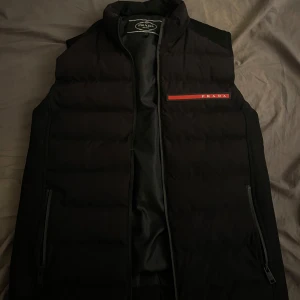 Svart dunväst från Prada - Snygg svart dunväst från Prada med diskret logga på bröstet i rött och vitt. Västen har hög krage, två dragkedjefickor och quiltad design. Perfekt för lager-på-lager och ger en clean och exklusiv look.