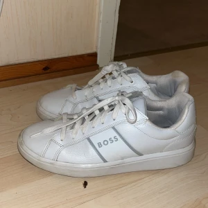 Vita sneakers från BOSS - Fräscha vita sneakers från BOSS med klassisk design och diskreta grå detaljer. Skorna har snörning och är tillverkade i skinn med rund tå och platt sula. Perfekta för en clean och stilren look.