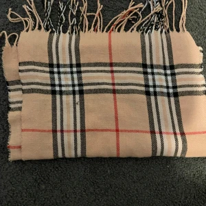 Beige rutig halsduk med fransar - Snygg beige halsduk med klassiskt rutigt mönster i svart, vitt och rött. Den har fransar i ändarna som ger en chill vibe och är perfekt att slänga över axlarna när det är kallt. Materialet känns mjukt och skönt mot huden. Pris kan även diskuteras