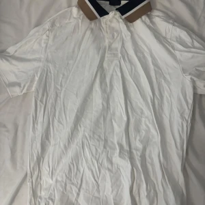 Vit BOSS t-shirt med kontrastkrage - Snygg vit t-shirt från BOSS med klassisk pikékrage i beige, svart och vit. Stor BOSS-logga på kragen ger en extra cool touch. T-shirten har korta ärmar och är gjord i mjuk bomull, perfekt för en clean och stilren look.