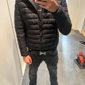 Svart pufferjacka med huva - Säljer en svart pufferjacka med huva och dragkedja framtill. Jackan har quiltad design och är lätt vadderad, perfekt för kyliga dagar. Passformen är normal och jackan har två fickor med dragkedja. Enkel och stilren look som funkar till det mesta.