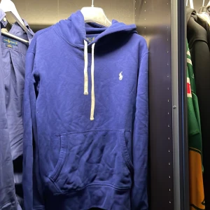 Blå hoodie från Polo Ralph Lauren - Säljer en blå hoodie från Polo Ralph Lauren med vit dragsko och klassisk logga på bröstet. Tröjan har känguruficka och huva, perfekt för en chill och stilren look. Materialet är mjukt och skönt, passar dig som gillar casual streetstyle.