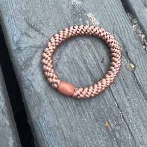Brunt flätat ringarmband i syntet - Snyggt brunt armband med flätad design, tillverkat i syntetmaterial. Rund form med en liten brun pärldetalj som accent. Perfekt för dig som gillar enkel och cool stil.