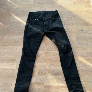 Nudie jeans - Säljer ett par svarta skinny jeans från Nudie med klassisk femficksdesign och snygga detaljer på bakfickorna. Jeansen har låg midja och är tillverkade i stretchig bomullsdenim för en tight passform. Perfekta för dig som gillar en slimmad look.